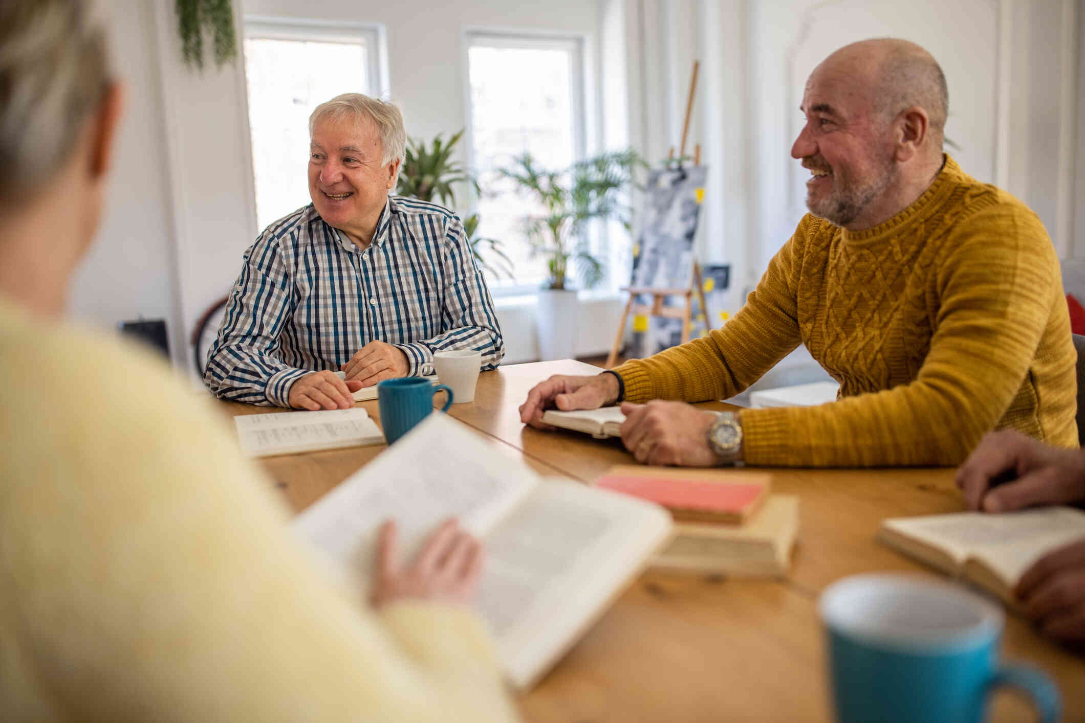 A group of mature men sit around a table and smile with books open and coffee mugs in front of them.